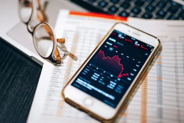 Smartphone displaying a financial chart beside glasses, paperwork, and a laptop.
