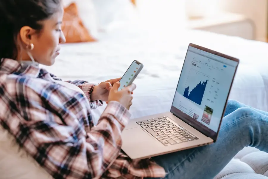 dba-investment-criteria-banner.webp Woman using a smartphone beside a laptop displaying a financial chart.