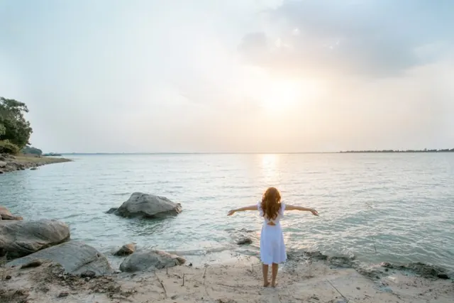 Woman standing at the water’s edge with outstretched arms at sunset.