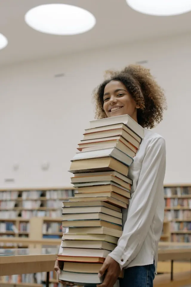 Smiling woman holding a tall stack of books in a library.